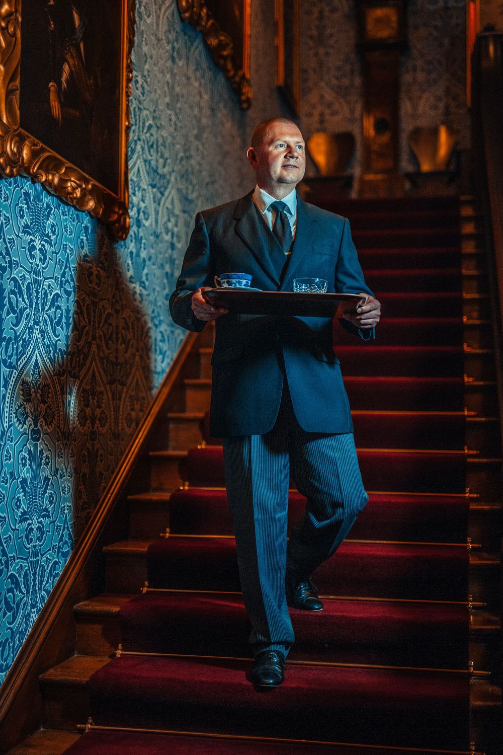 A businessman in a dark suit sits on ornate carpeted stairs in dramatic lighting with patterned wallpaper behind him.