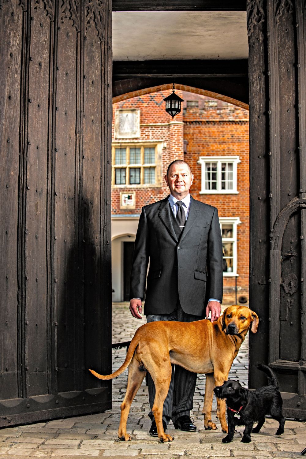 Two people in formal attire stand with a brown dog in front of a historic brick building entrance.