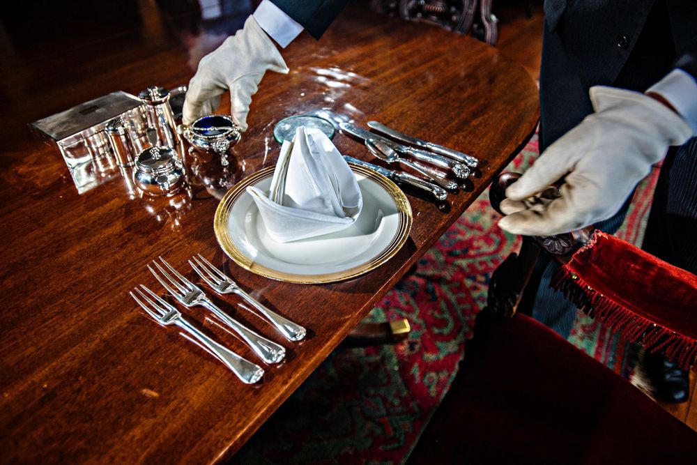 A formal place setting on a wooden table with silver utensils and a folded napkin being arranged by gloved hands.
