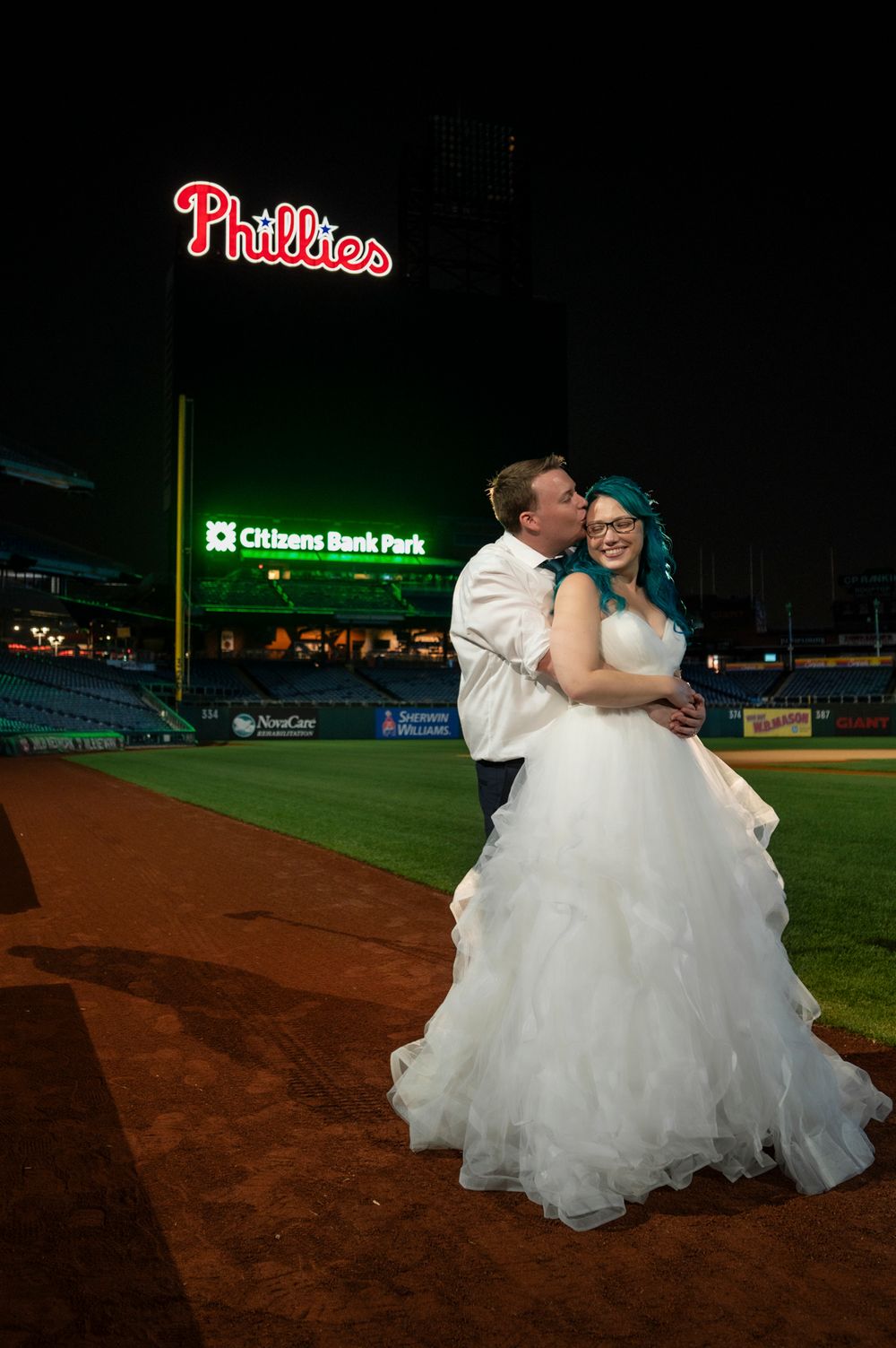 A couple in wedding attire embraces on a baseball field at Citizens Bank Park at night with stadium lights glowing.
