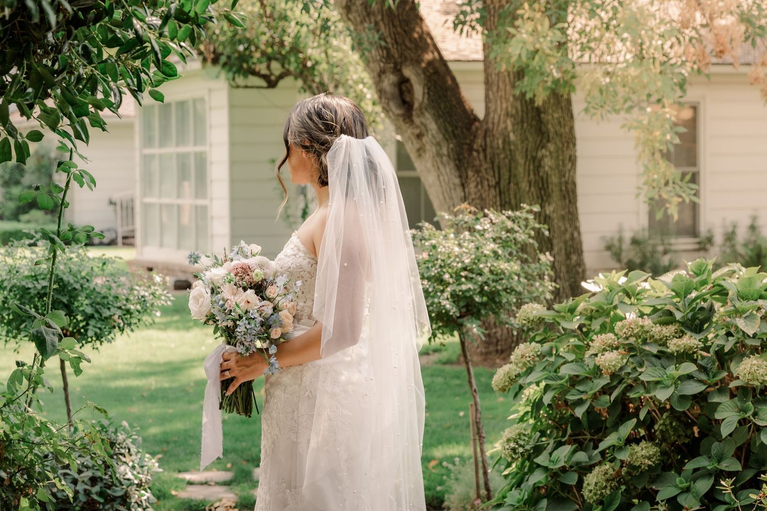 A bride in a flowing veil holds a pastel bouquet in a garden setting.