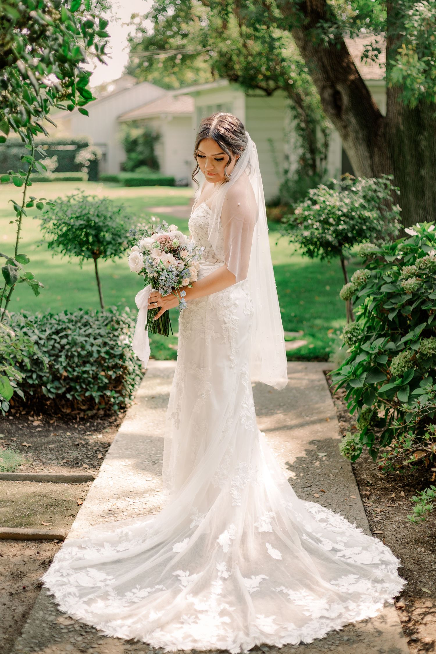 A bride in a lace gown with long train poses with bouquet in a garden pathway.