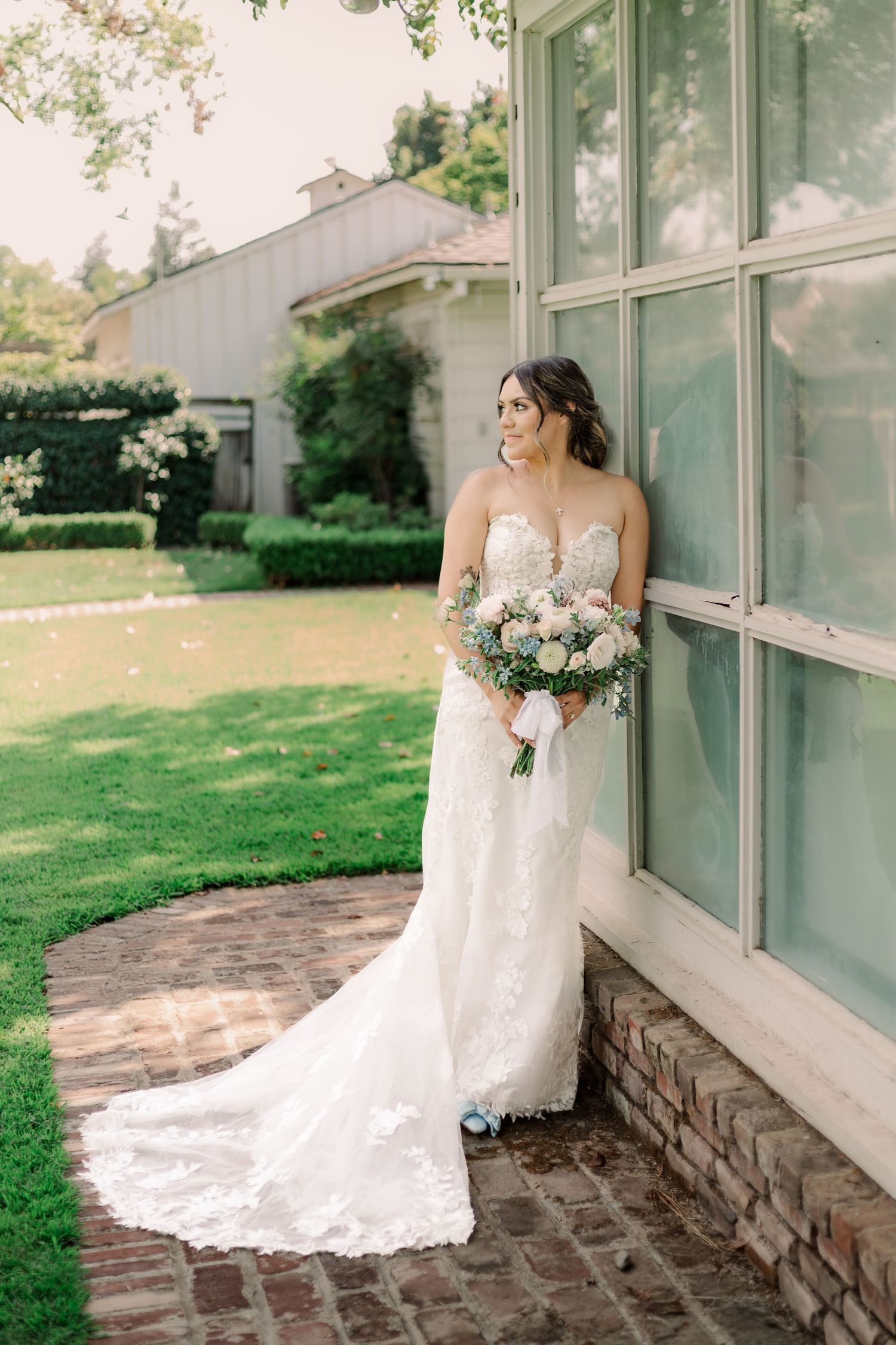 A bride in a lace wedding gown holds a pastel bouquet while standing beside large windows on a brick patio.