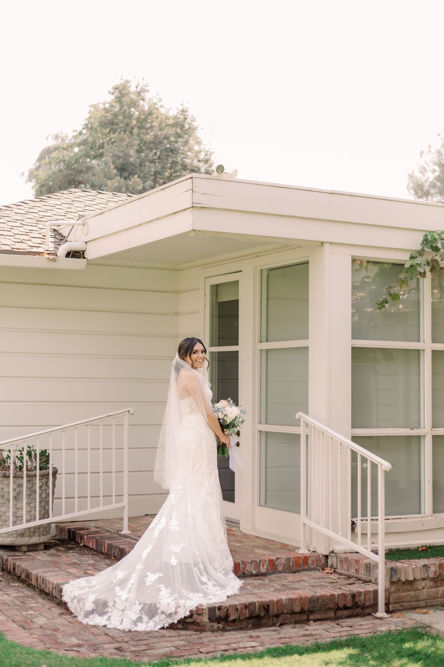 A bride in a flowing lace gown poses on brick steps outside a white house with large windows.