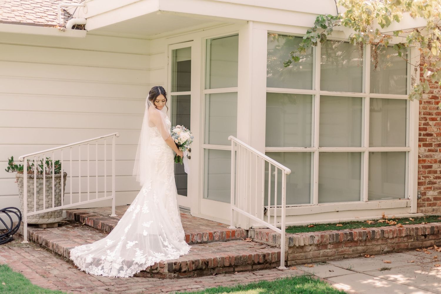 A bride stands on brick steps wearing a long-train lace wedding dress beside a white railing.
