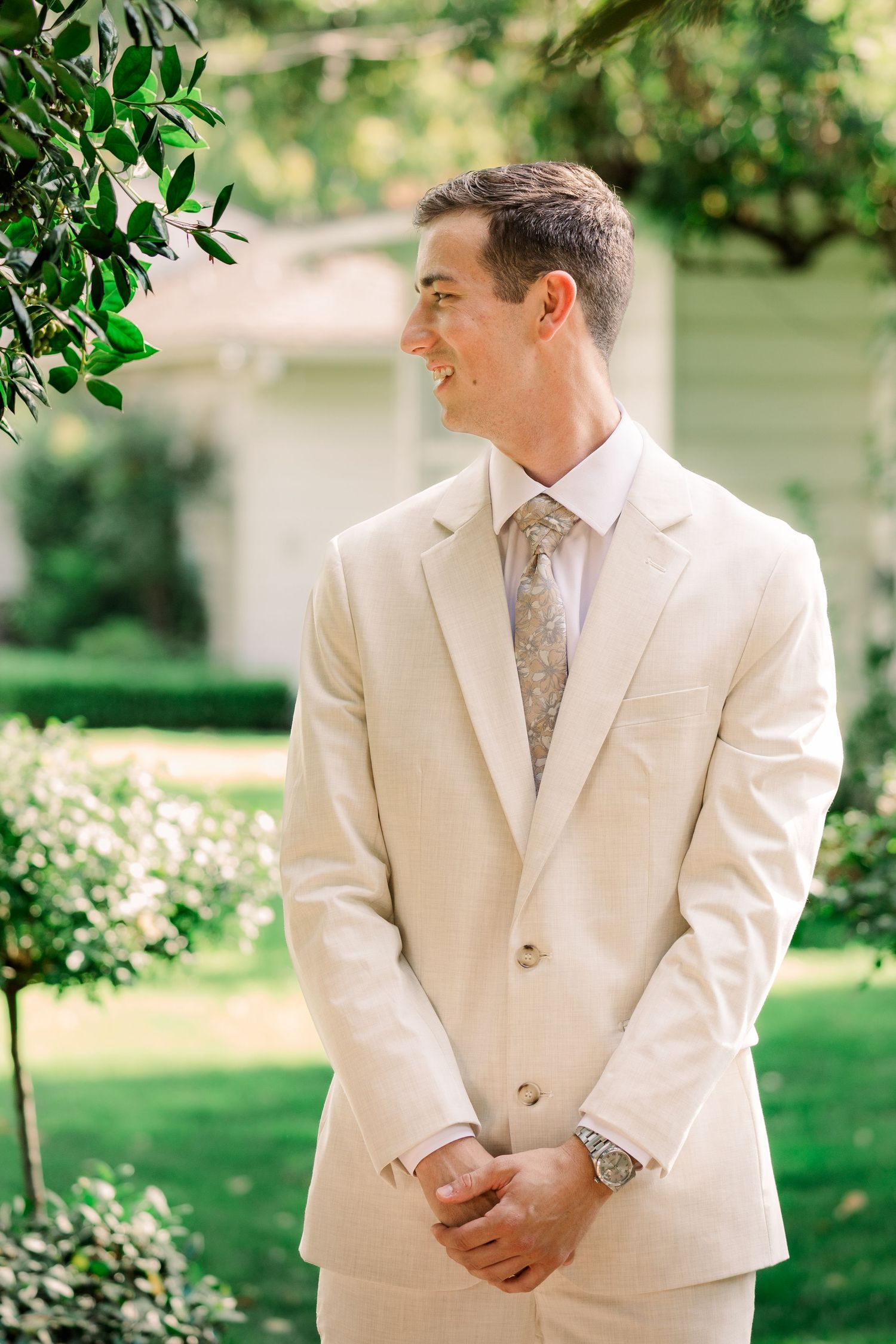 A groom in a light cream suit and patterned tie smiles while standing in a garden setting.