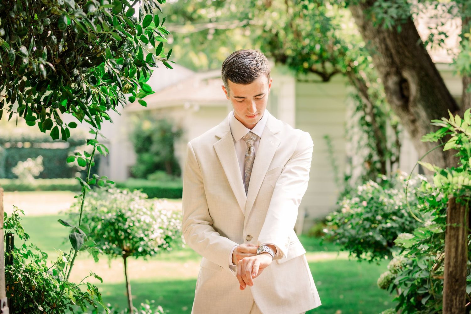 A groom in a cream colored suit checks his watch while standing in a garden.