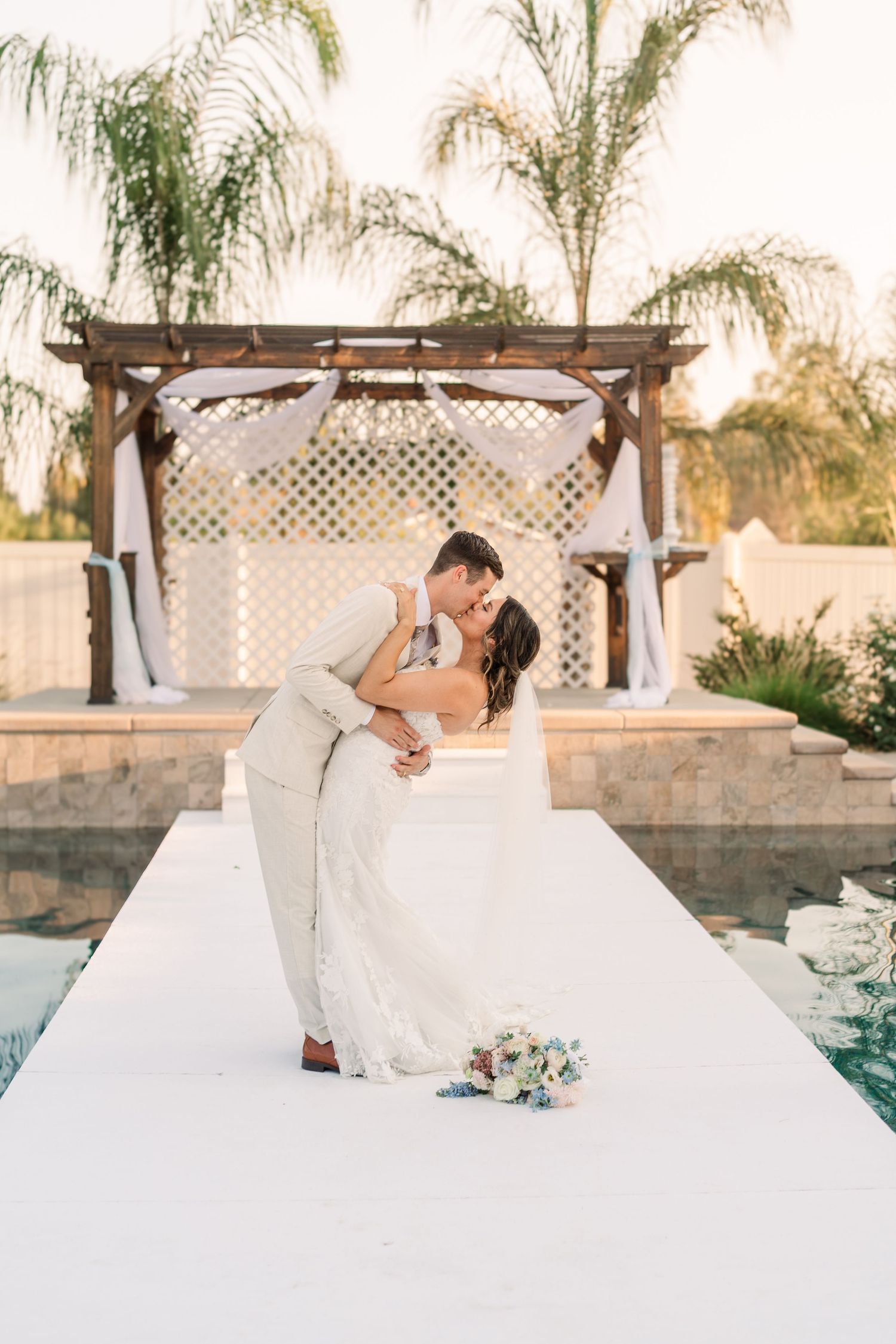 Newlyweds share a romantic dip kiss on a white platform near a pool with palm trees in the background.