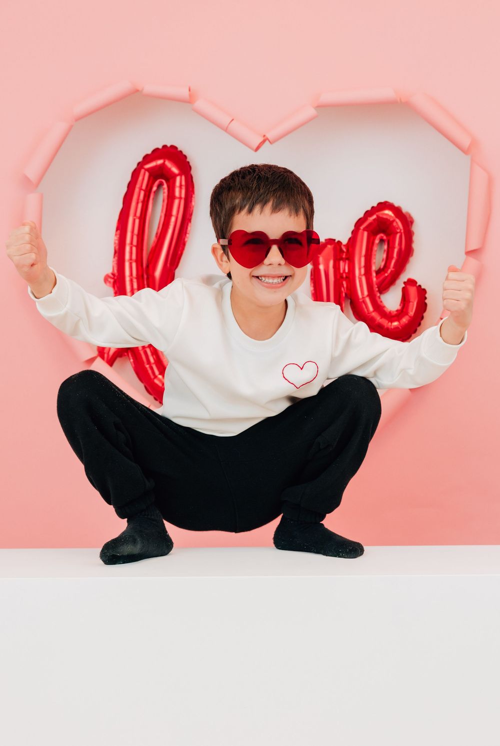 Smiling boy in heart sunglasses squats in front of a pink heart with red love balloons.