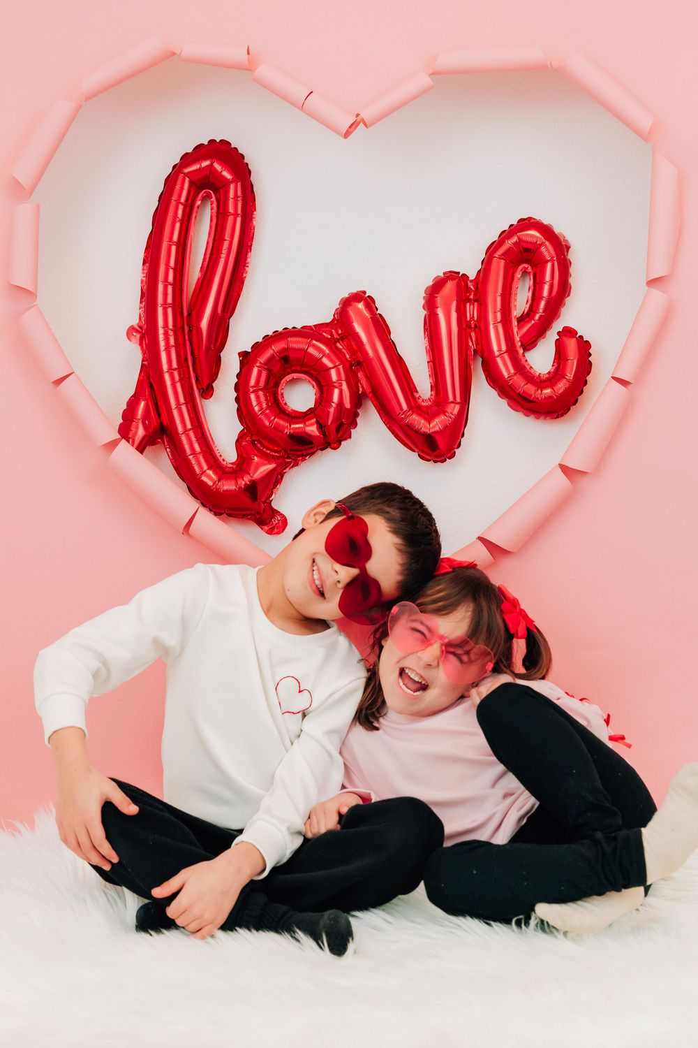 Two kids in heart sunglasses sit laughing together in front of a red love balloon display.