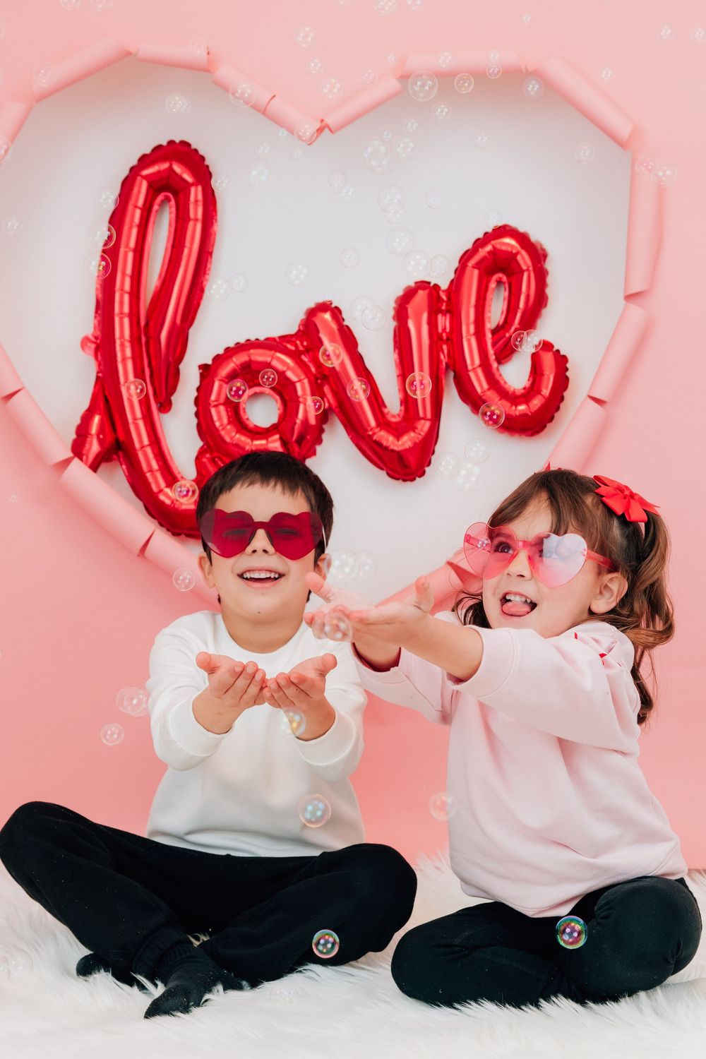Two kids in Valentine's outfits sit on fur rug tossing confetti before a pink heart love backdrop.
