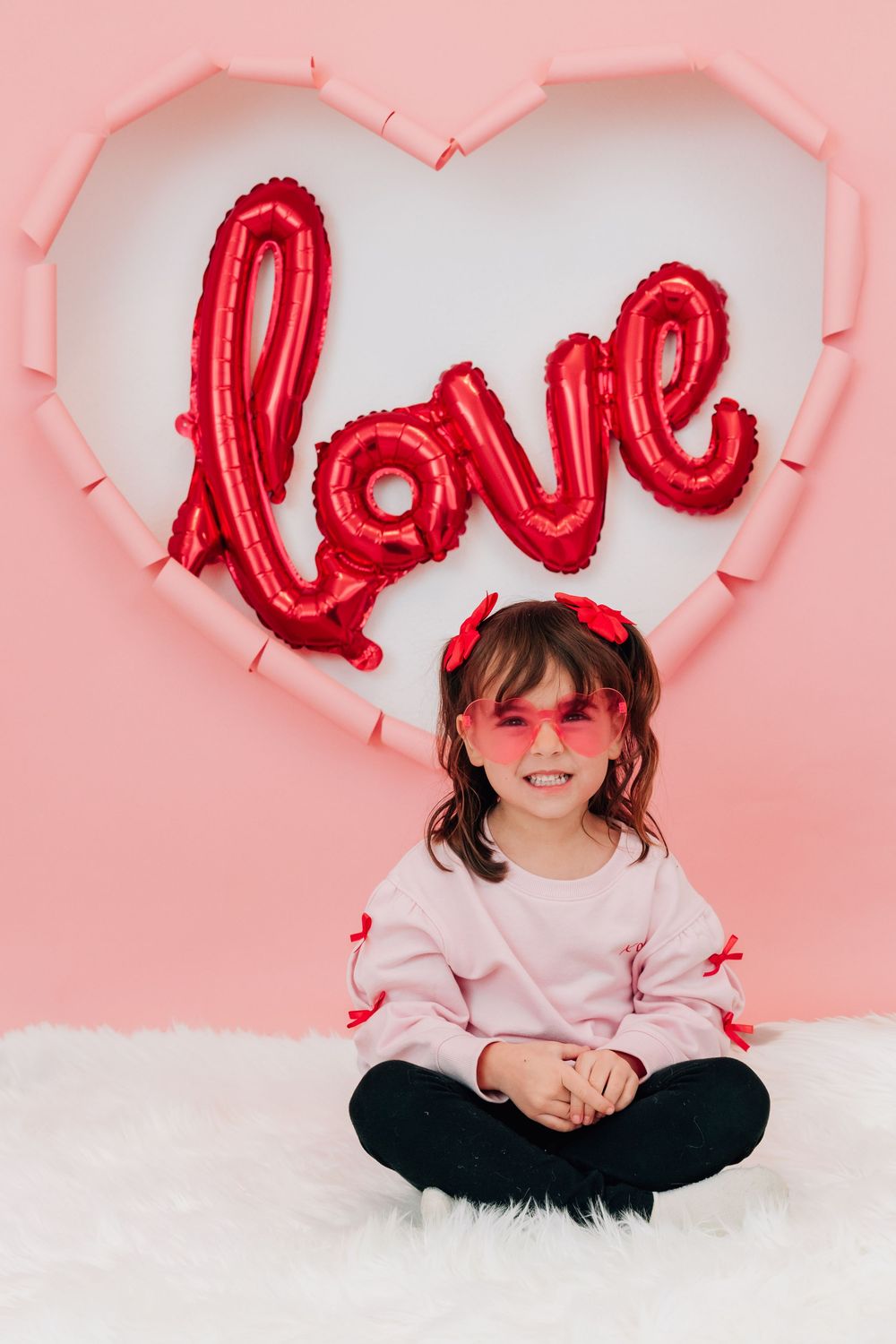 A smiling girl in pink sits cross-legged on fur rug before a heart backdrop with red love balloon.