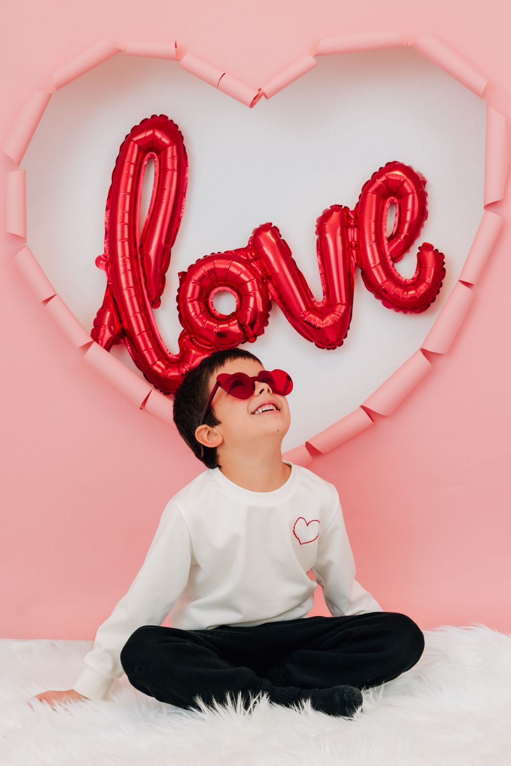 A boy in white sweatshirt sits cross-legged looking up at a red love balloon on pink heart backdrop.
