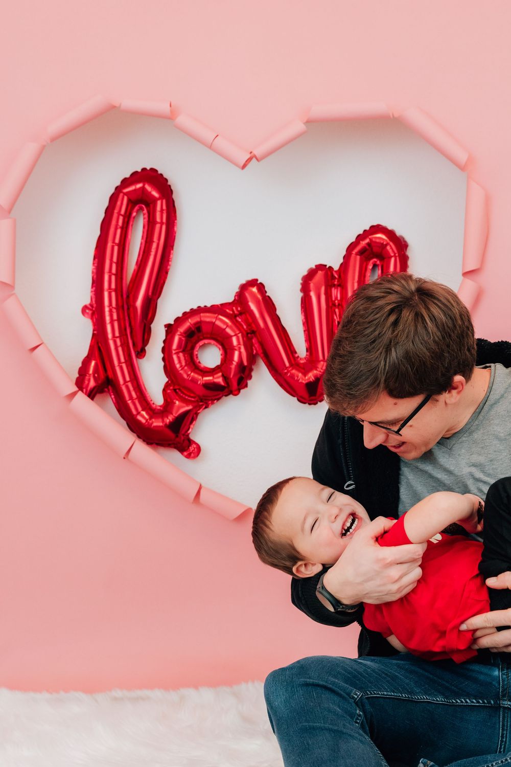 A father lovingly gazes at his baby in red outfit before a pink heart wall with love balloon.