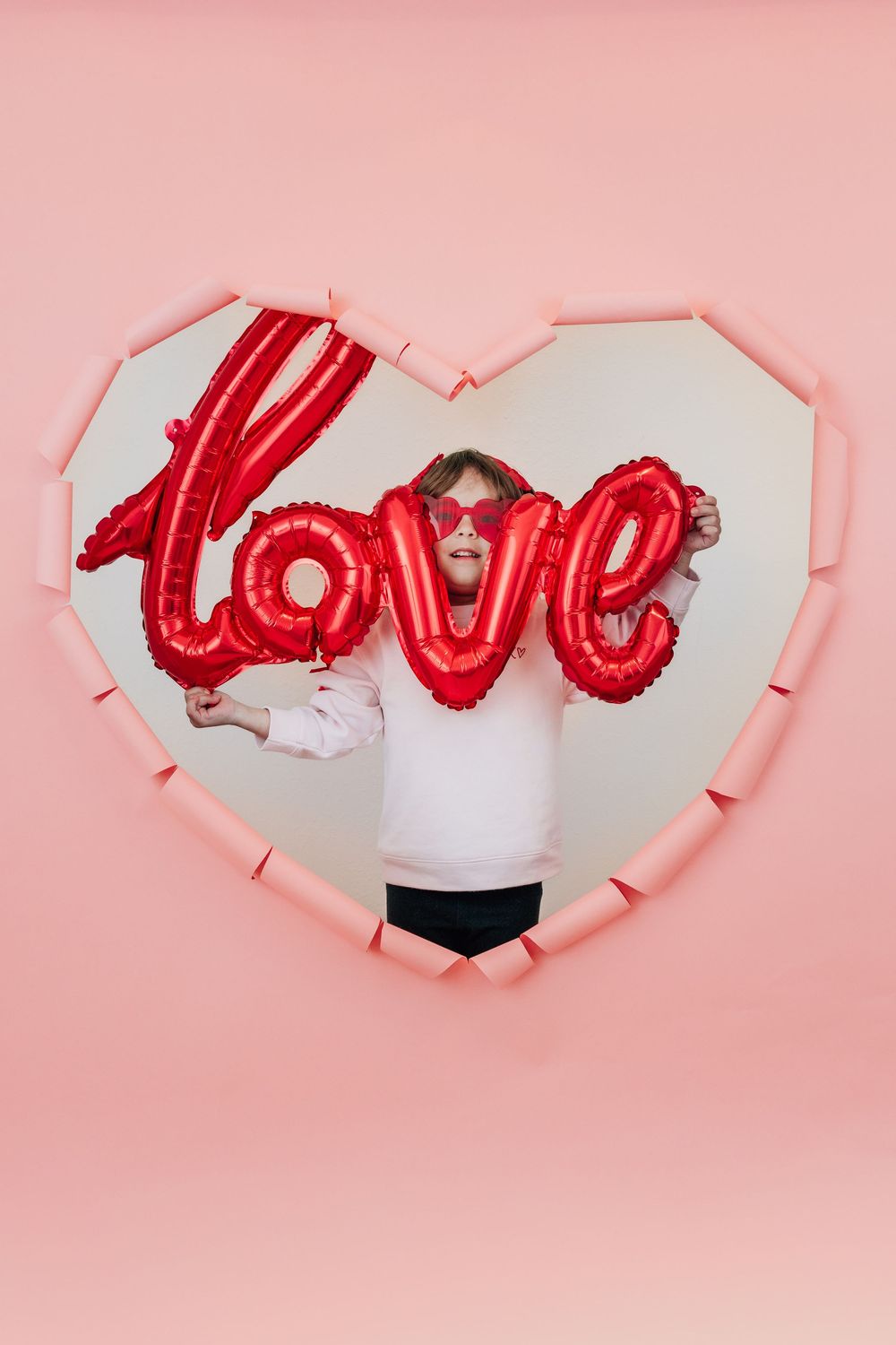 A girl in pink holds a red love foil balloon while standing inside a heart-shaped pink wall cutout.