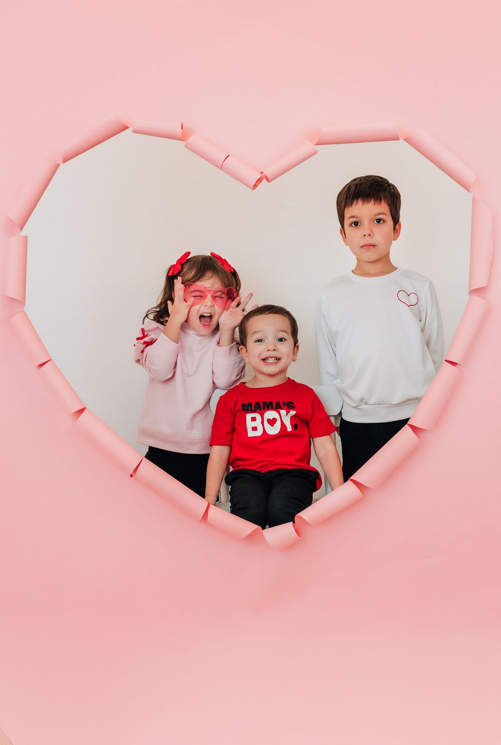 Three siblings pose together inside a heart-shaped pink wall cutout for a Valentine's Day photo.
