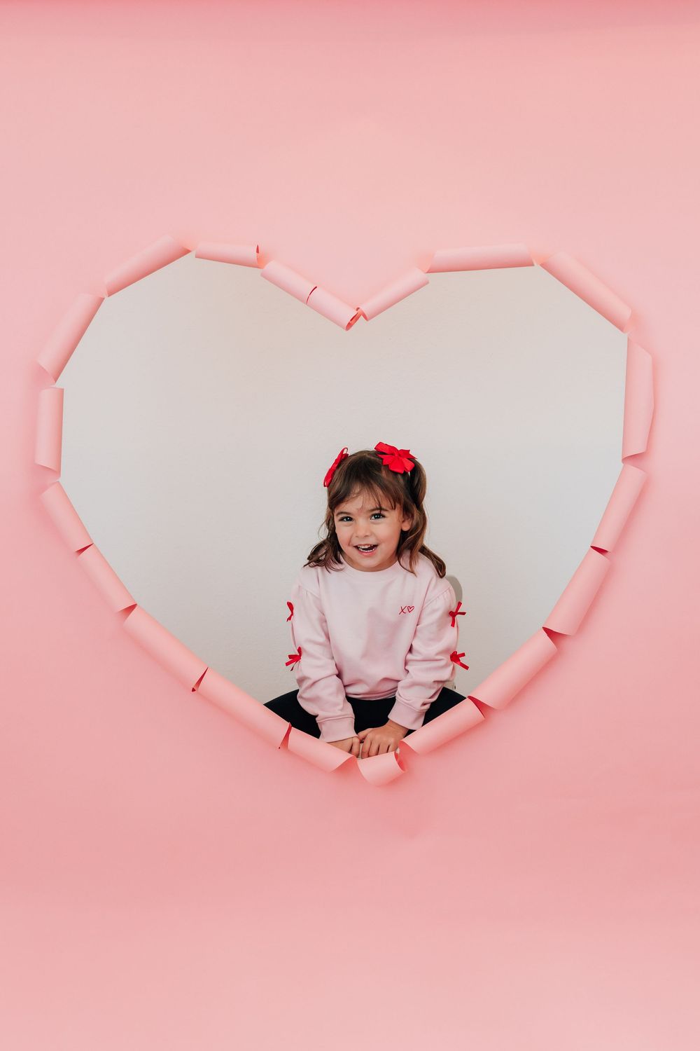 A cheerful girl in pink with a red bow sits inside a heart-shaped pink wall cutout smiling.