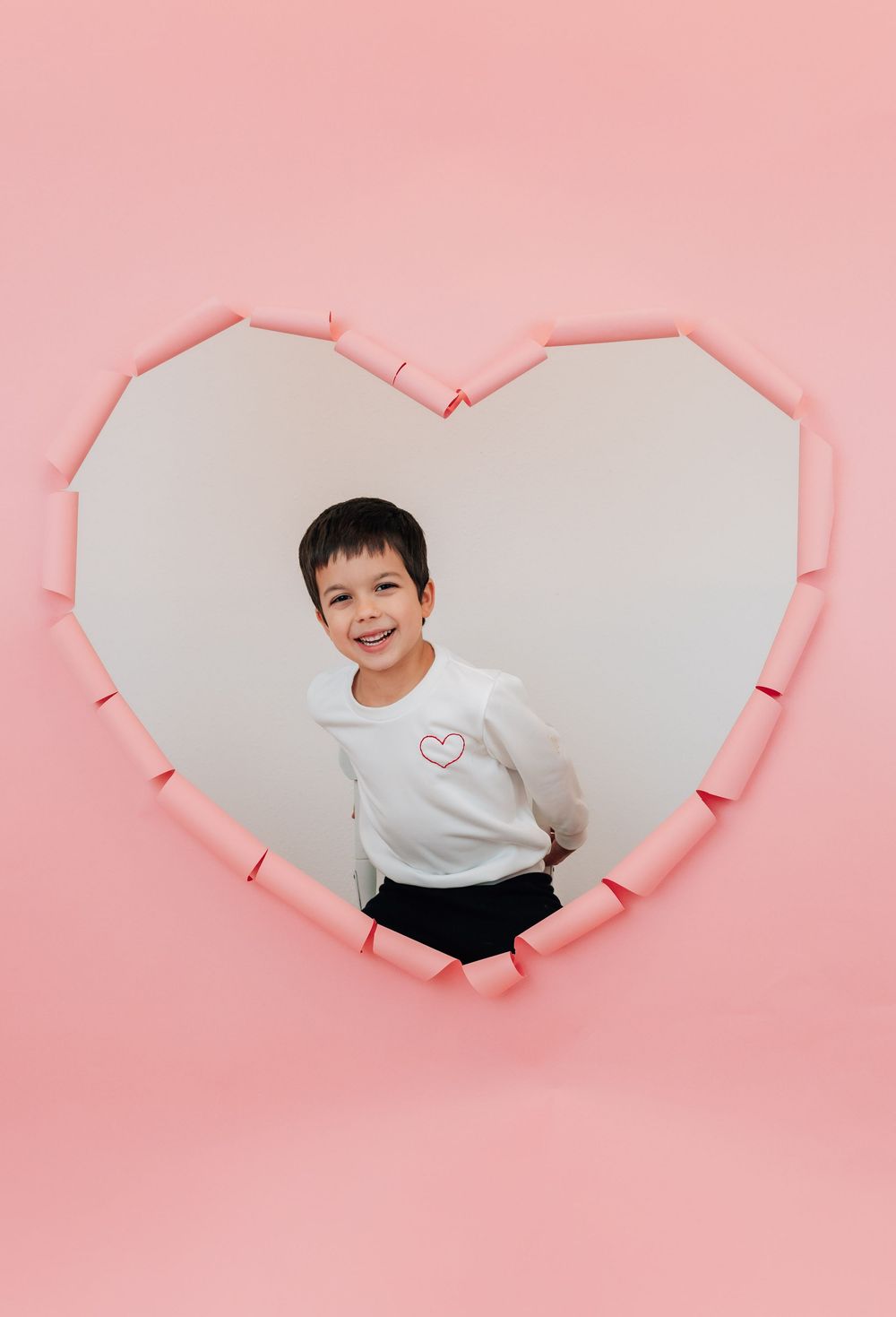 A smiling boy in white heart sweatshirt poses inside a heart-shaped pink wall cutout backdrop.