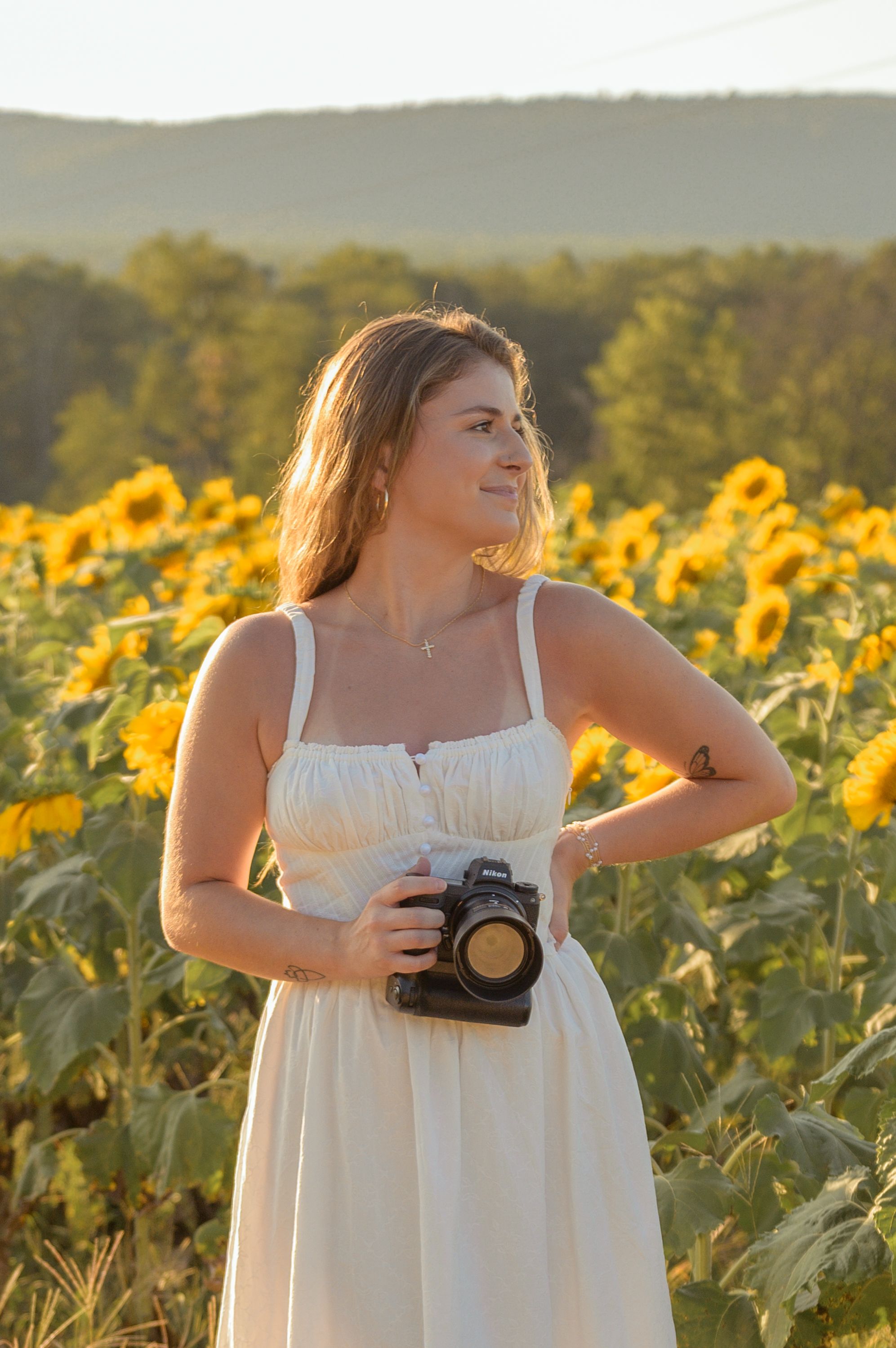 A photographer in a flowing white dress captures images in a vibrant sunflower field at golden hour.
