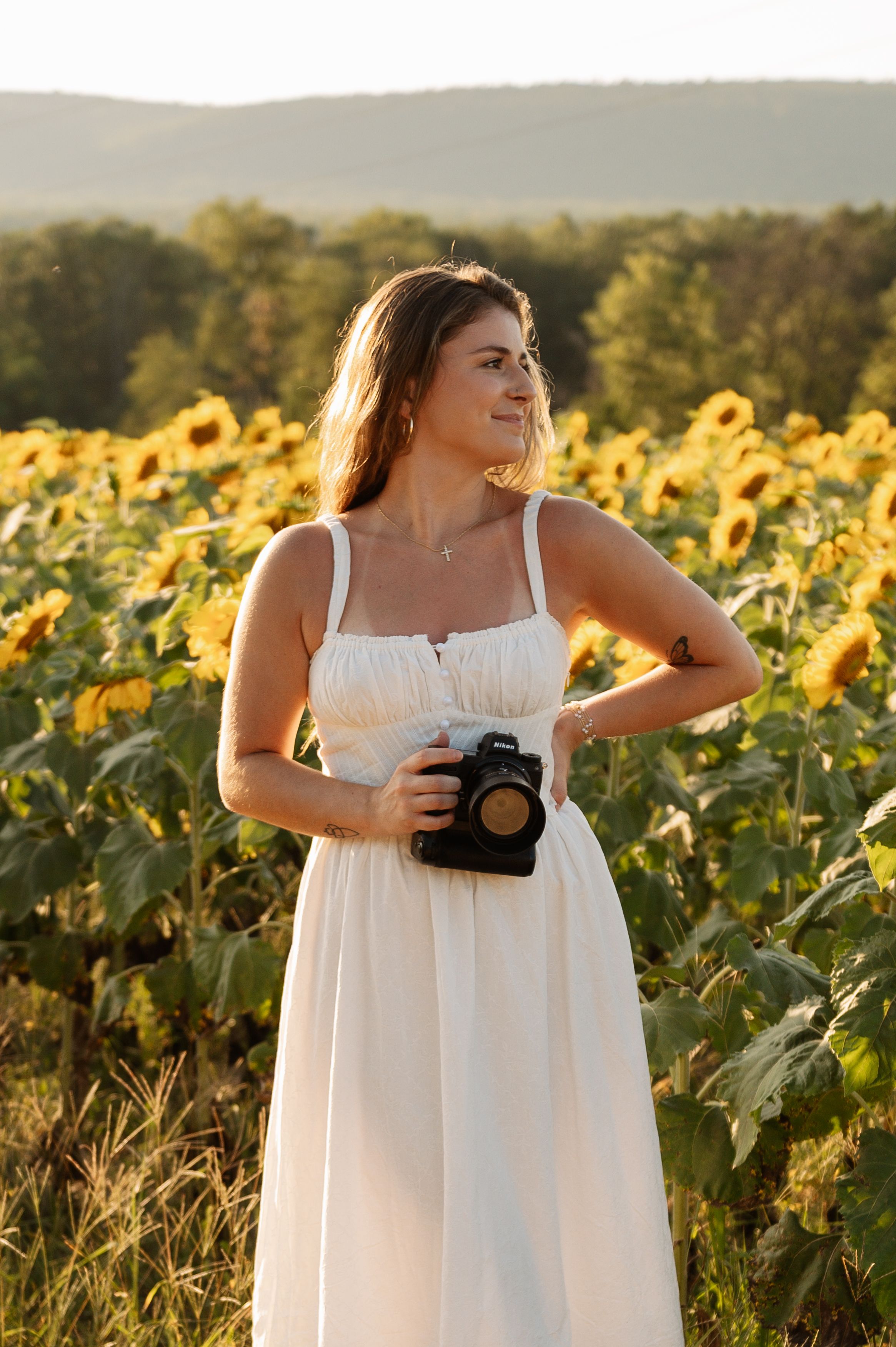 A photographer in a flowing white dress captures images in a vibrant sunflower field at golden hour.