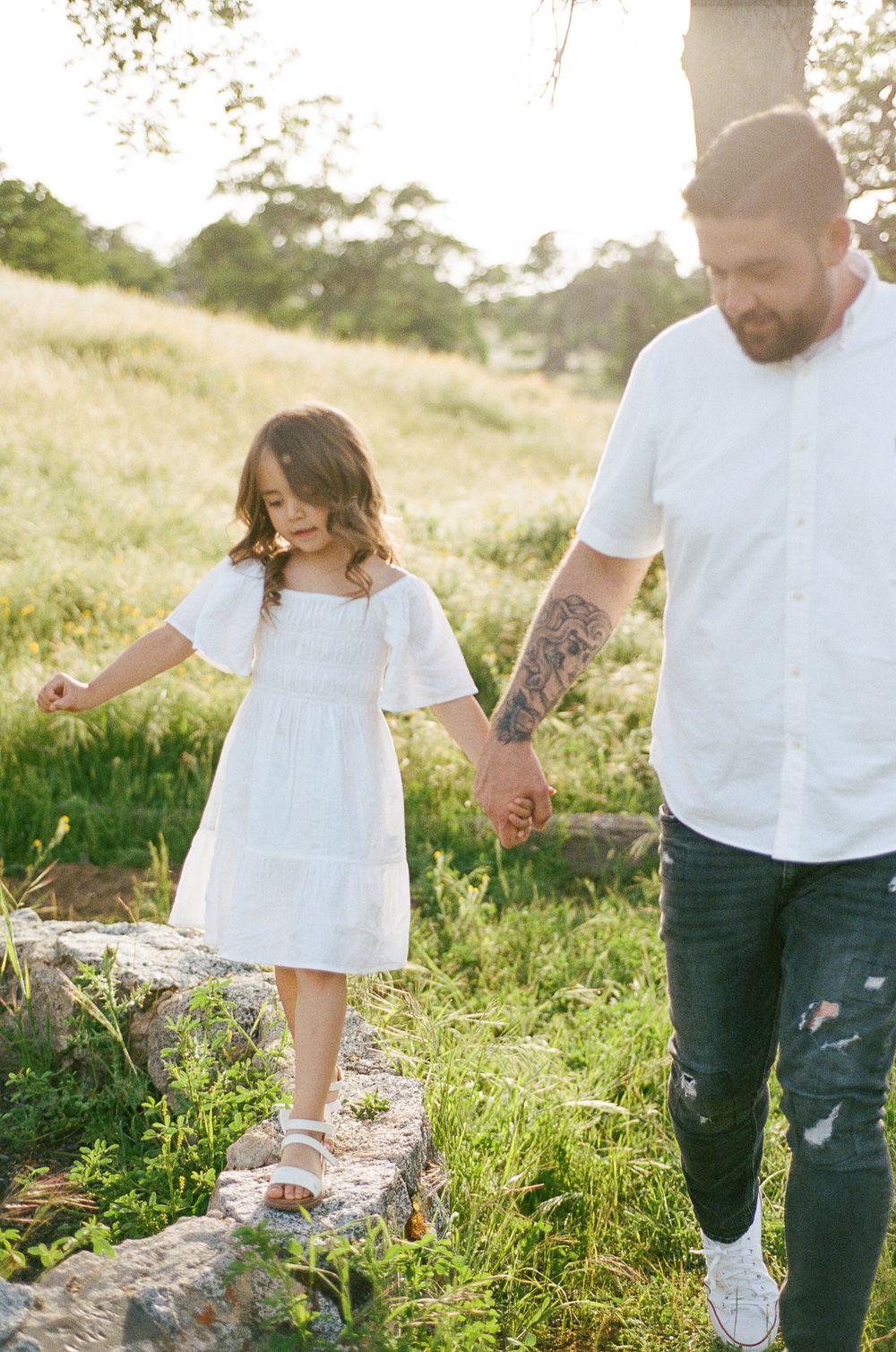 A couple walks hand in hand through a sunlit meadow wearing white tops and jeans during a romantic outdoor photoshoot.
