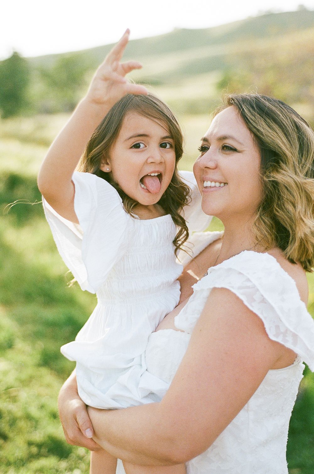Mother and child share playful moments together outdoors in white dresses during a sunny afternoon photoshoot.