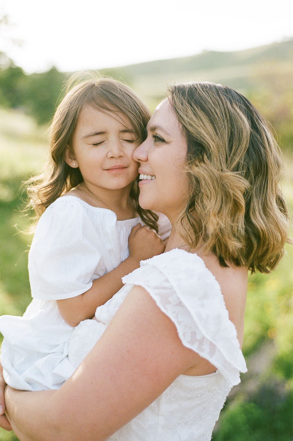 A tender moment captured outdoors in natural sunlight as a parent and child share an embrace while wearing white clothing.
