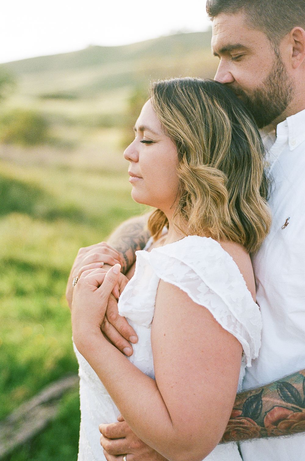 Couple embraces tenderly in a sunlit meadow wearing white clothing.
