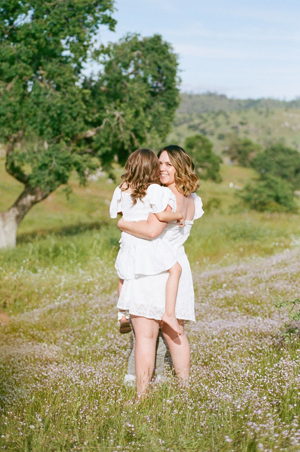 Two people in white dresses dance together in a wildflower meadow on a sunny afternoon.