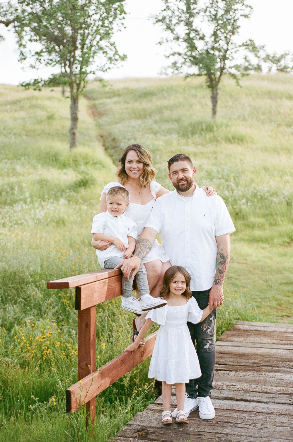 A family poses together on a wooden boardwalk surrounded by tall grass and trees in a natural outdoor setting.