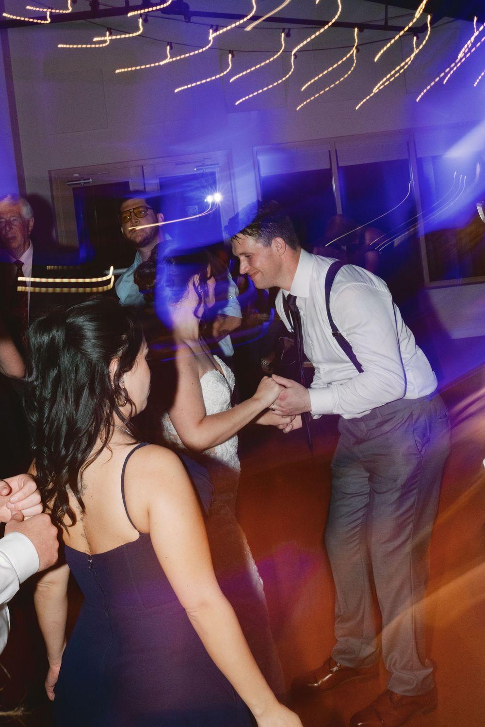 People dancing at a wedding reception with purple mood lighting and string lights decorating the ceiling.