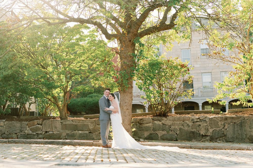 A couple in wedding attire embraces under lush green trees during an outdoor photoshoot at a historic stone venue.