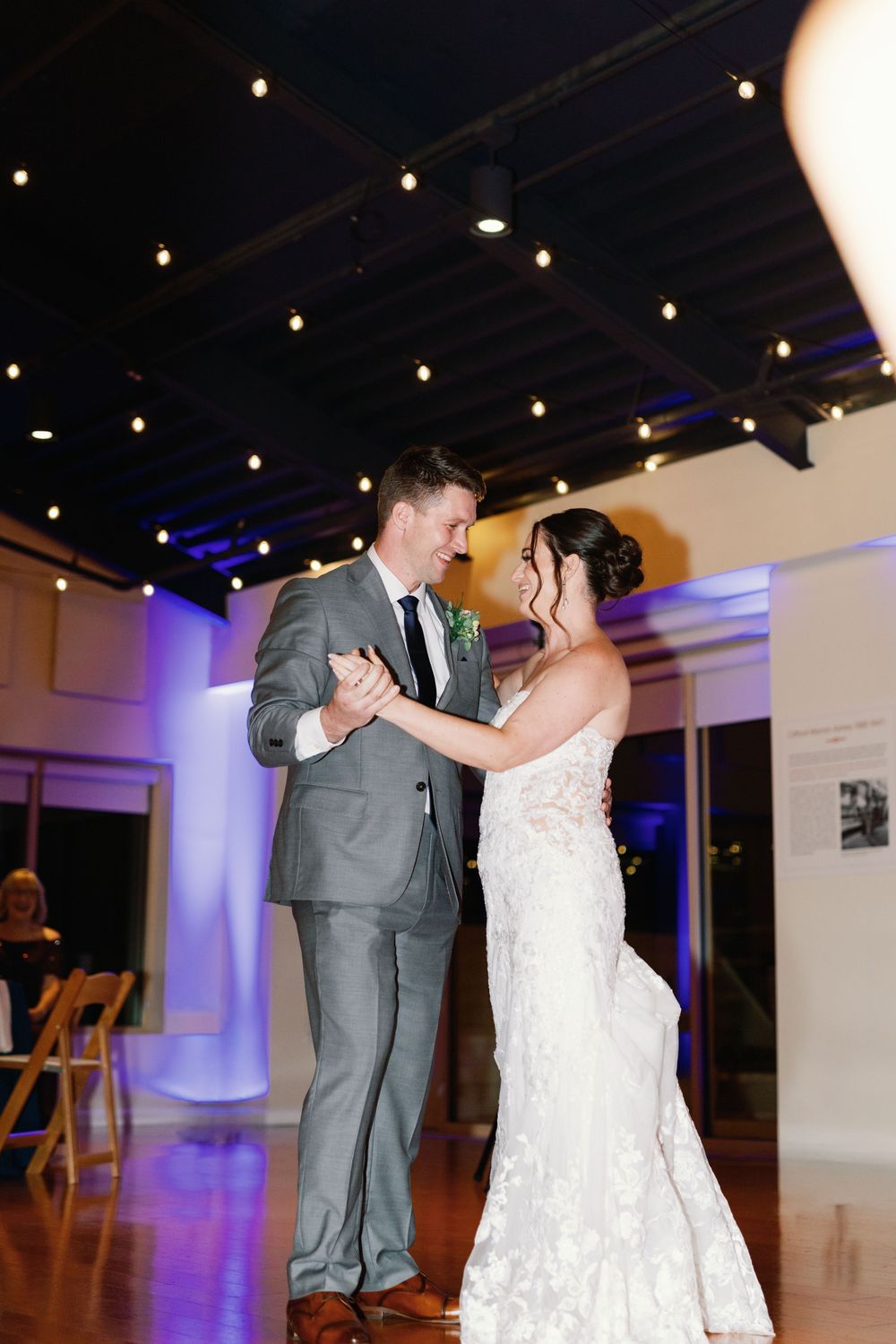 A couple shares their first dance at a wedding reception under string lights in an elegant white venue with blue uplighting.