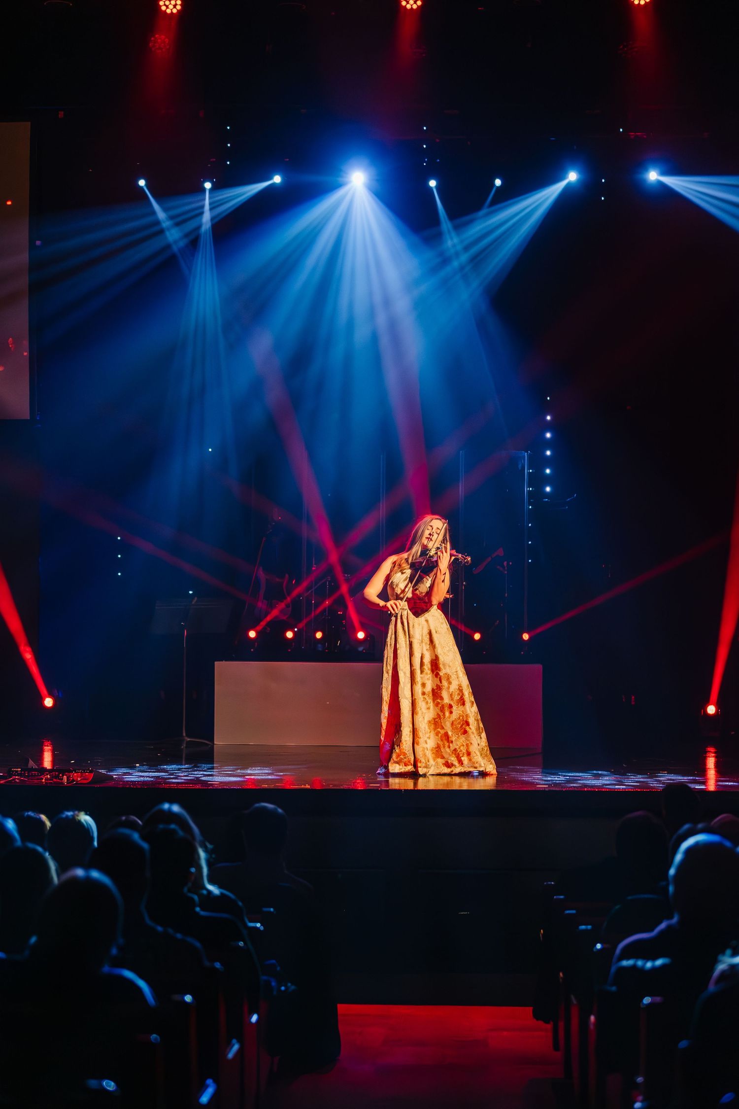 Dramatic stage performance with red and blue spotlights illuminating performers in golden costumes before a seated audience.