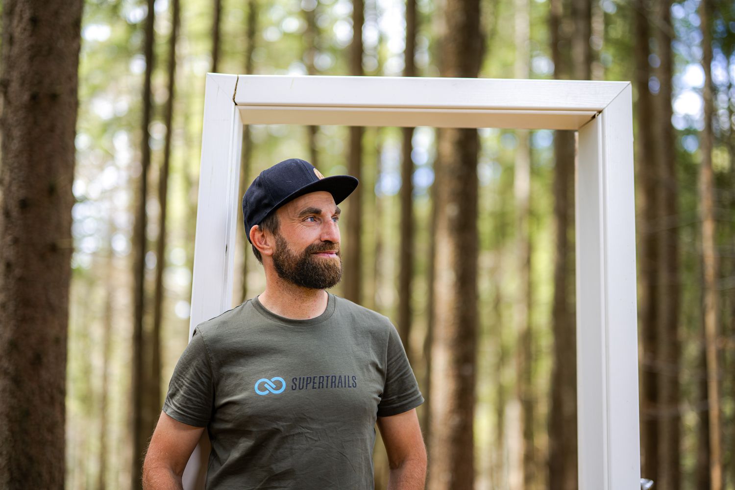 A bearded man in a black t-shirt and cap stands in a forest setting near a white door frame.