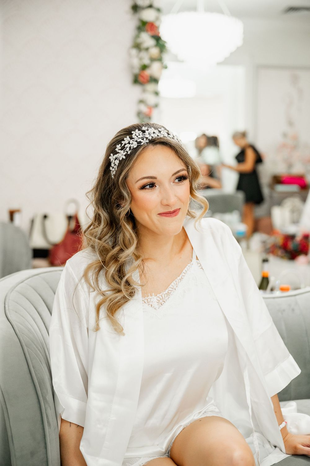 Person in white robe and tiara getting ready in a bright room with floral decorations.
