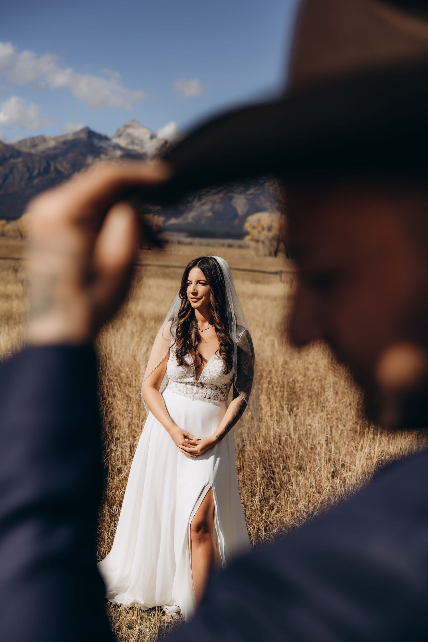 A bride in a white dress stands in a golden field with mountains in the background during a wedding photoshoot.