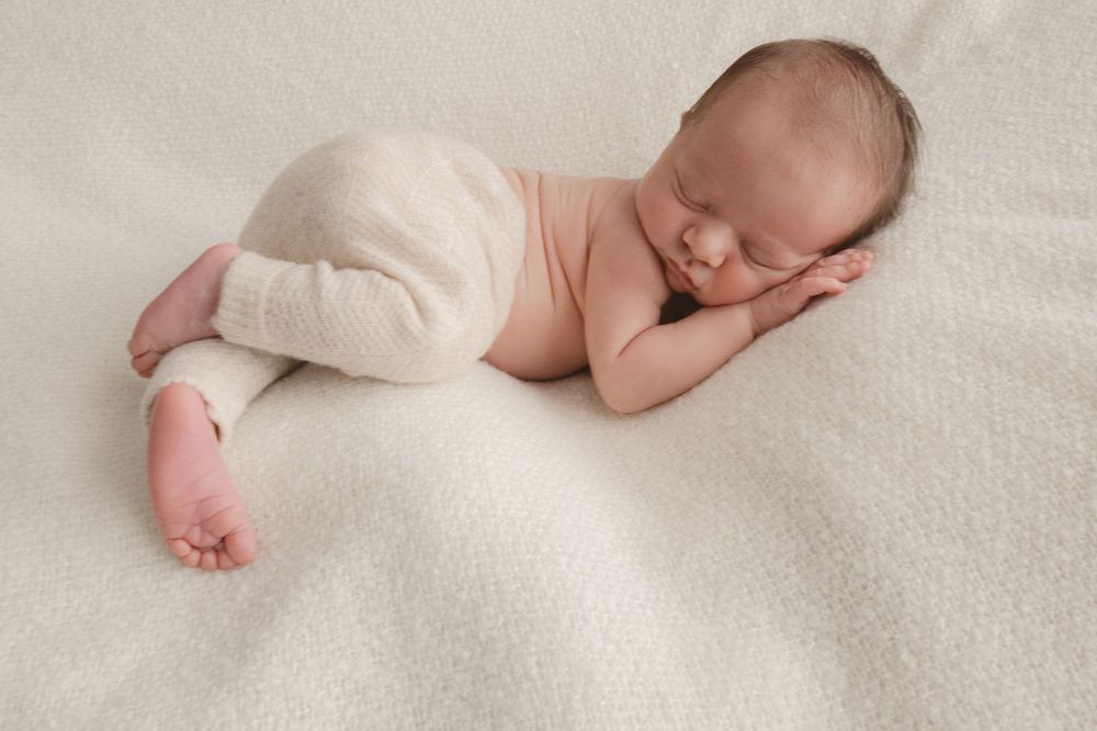 Peaceful newborn baby sleeping on white blanket wearing cream knit pants.