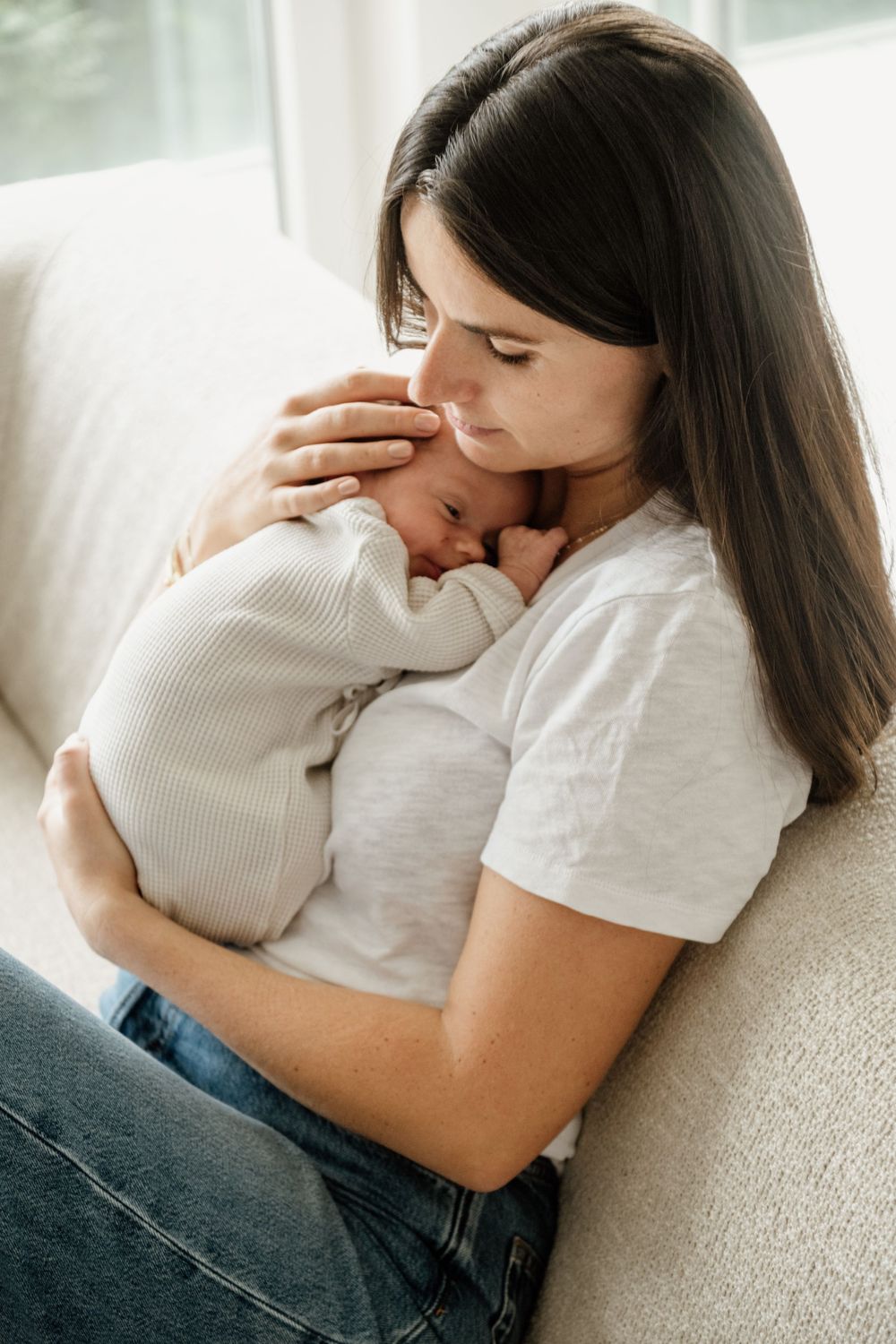 Mother tenderly holding and nuzzling her sleeping newborn baby while seated on a cream sofa.