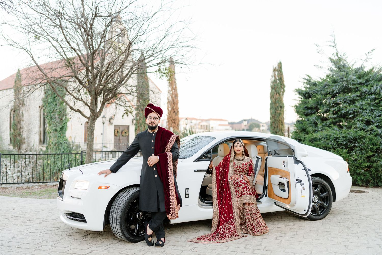 A couple in traditional wedding attire pose with a white luxury car outside a building with cypress trees.
