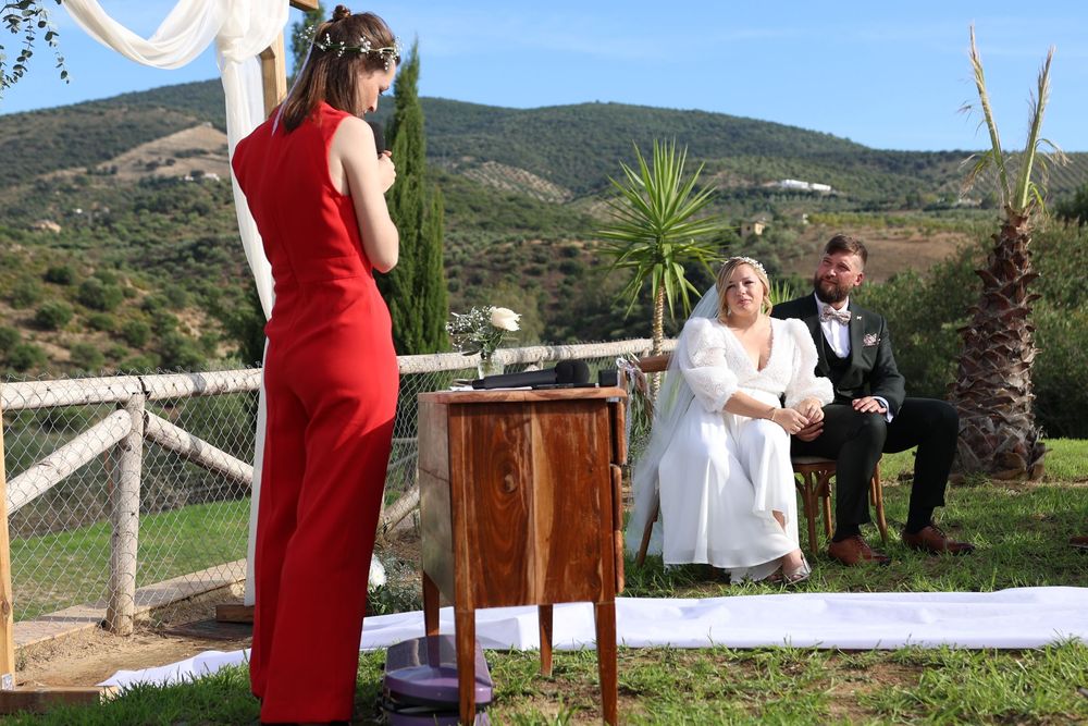 An outdoor wedding ceremony takes place on a hillside with a person in red officiating and palm trees in the background.