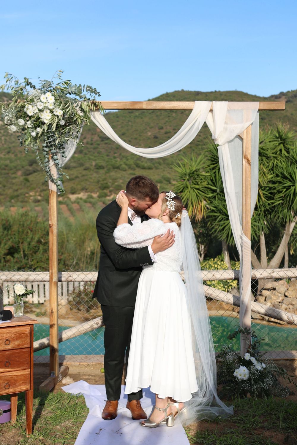A couple shares their first kiss at an outdoor wedding ceremony under a wooden arch draped with white fabric.