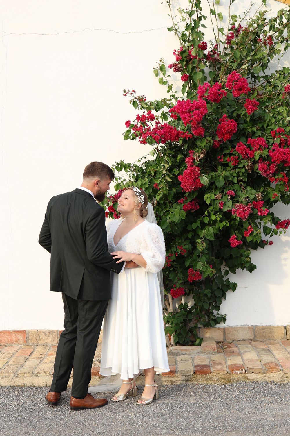 A couple shares a romantic moment against a white wall with vibrant red climbing roses in bloom.