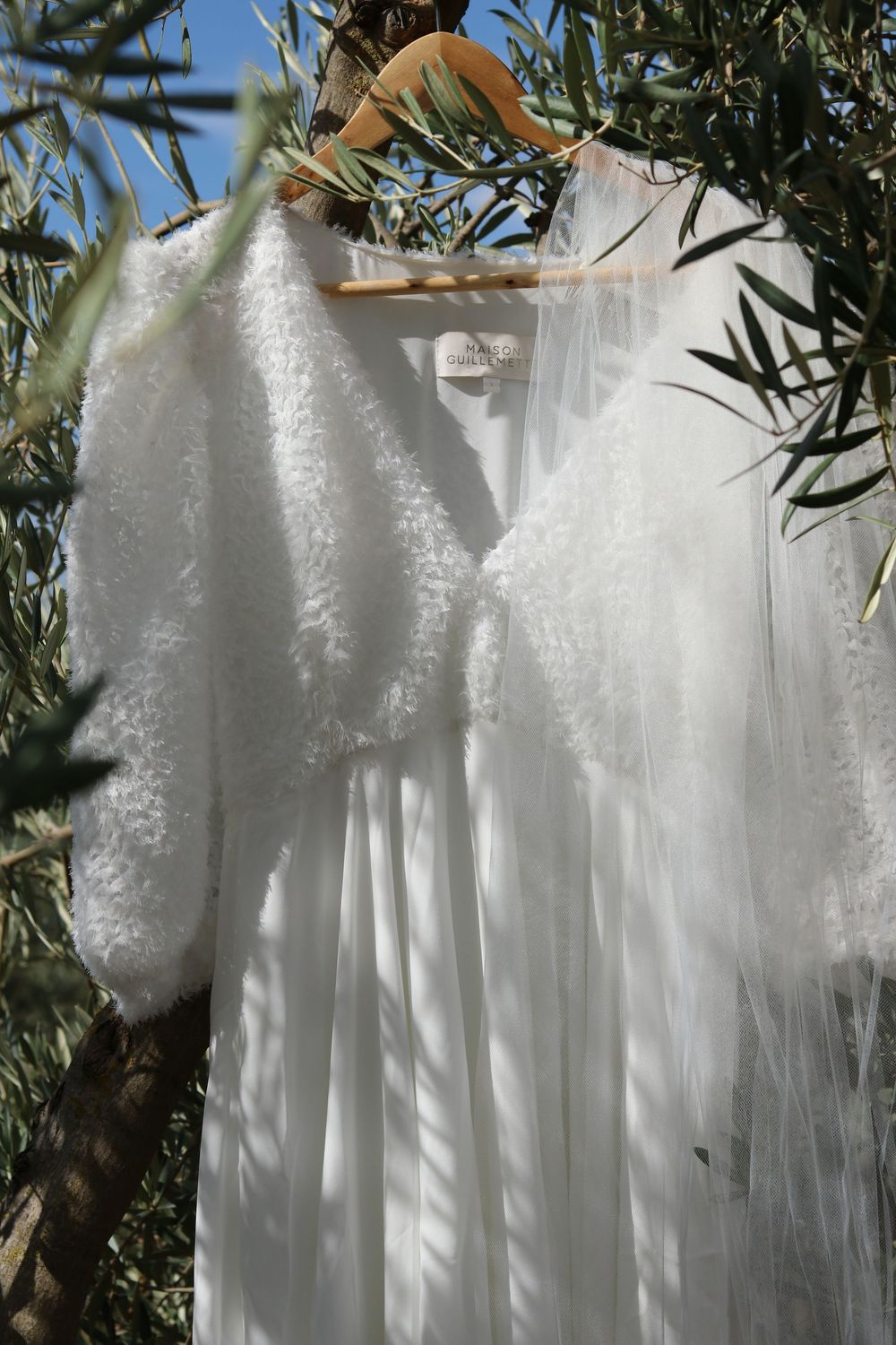 A delicate white lace wedding dress hangs on a wooden hanger among olive tree branches in natural sunlight.