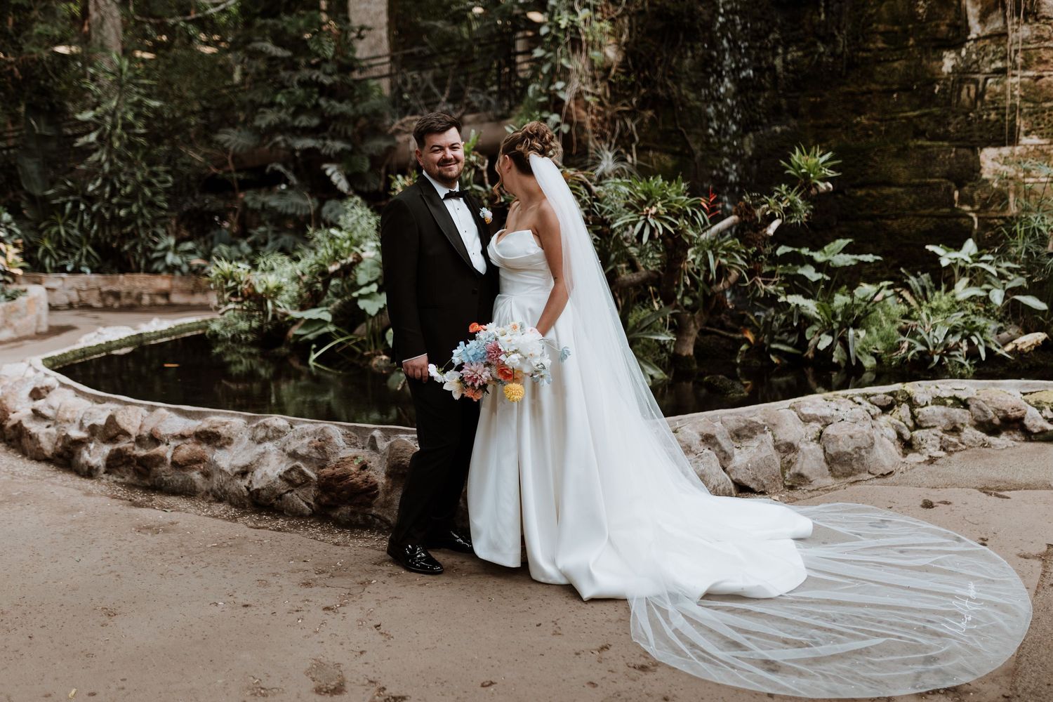 A couple in wedding attire stand together in the lush rain forest greenery at Chester Zoo.