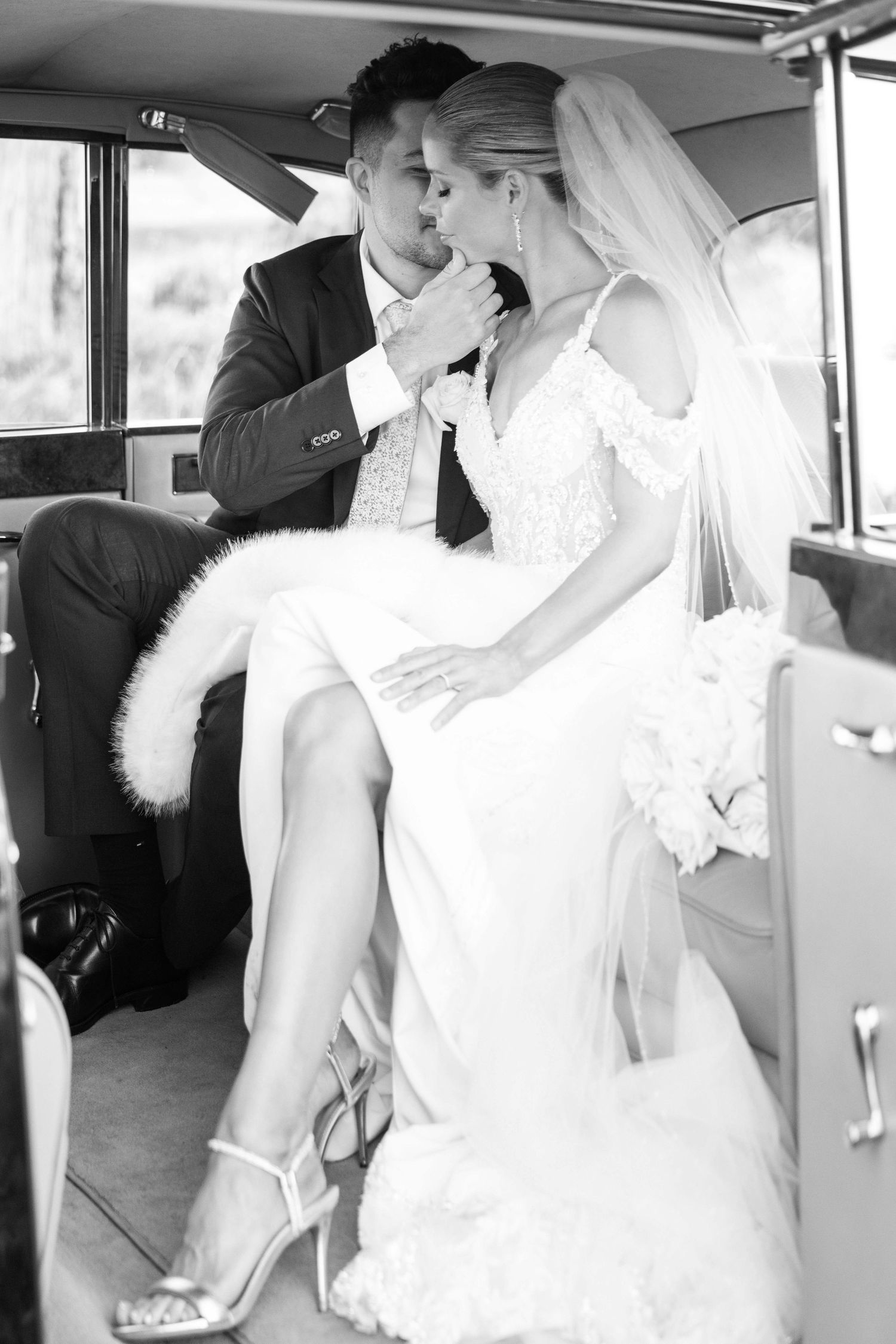 Bride and groom sharing intimate kiss inside vintage car, captured in elegant black and white.