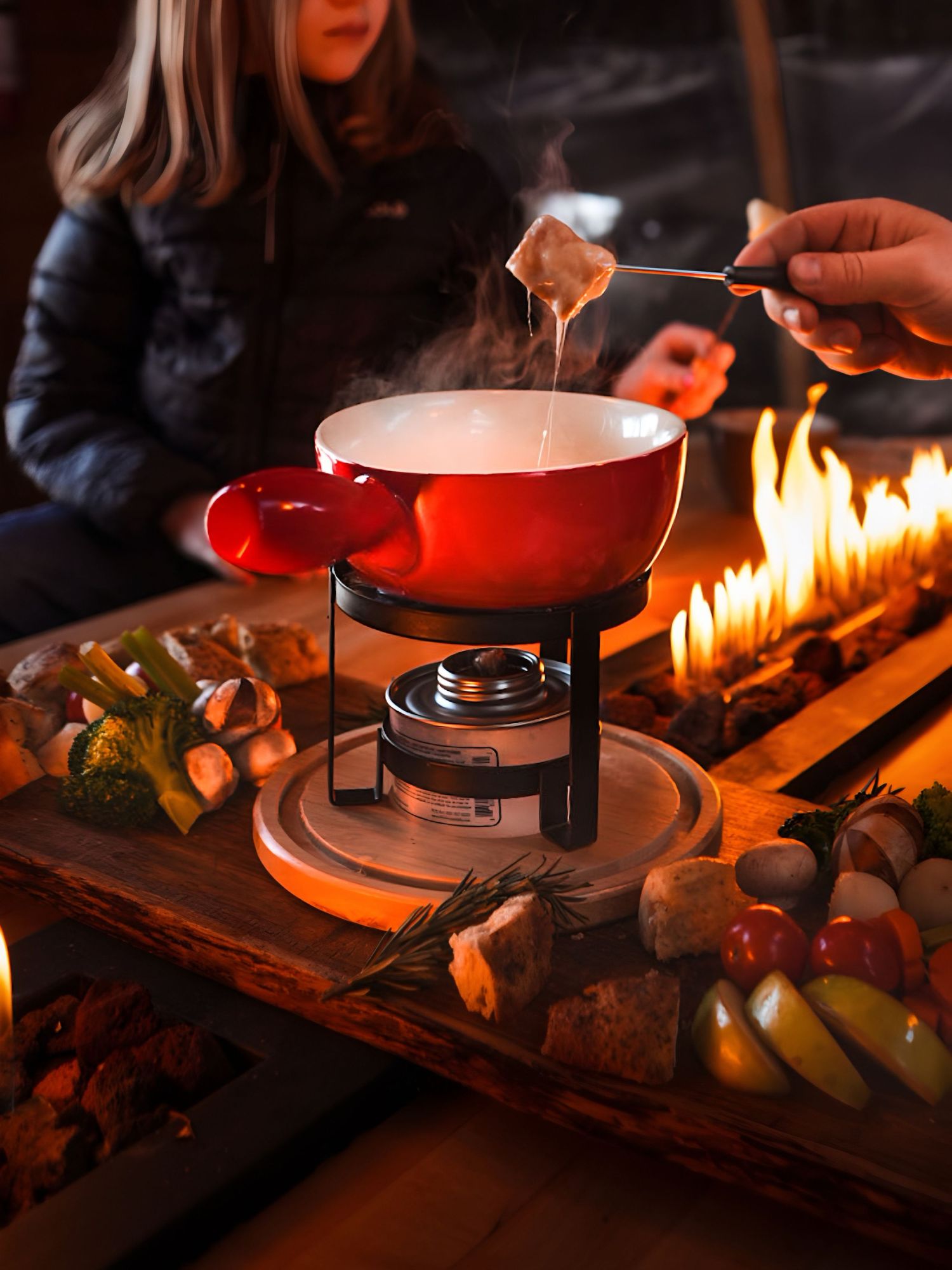 Cozy cheese fondue in red pot with flames and fresh ingredients on wooden table by firelight.