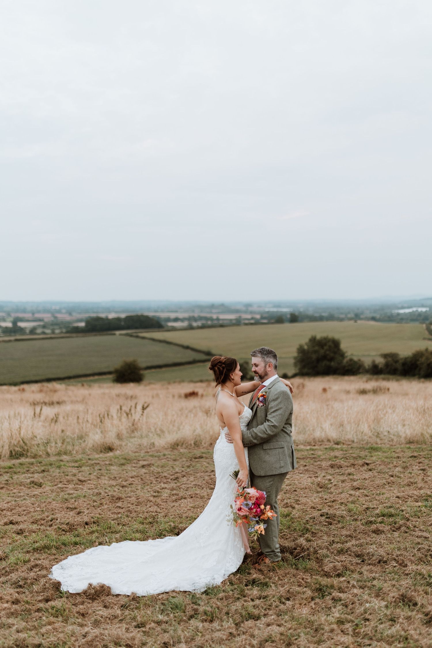 A couple shares a romantic moment on a hilltop overlooking rolling countryside fields with a beautiful bridal gown train.