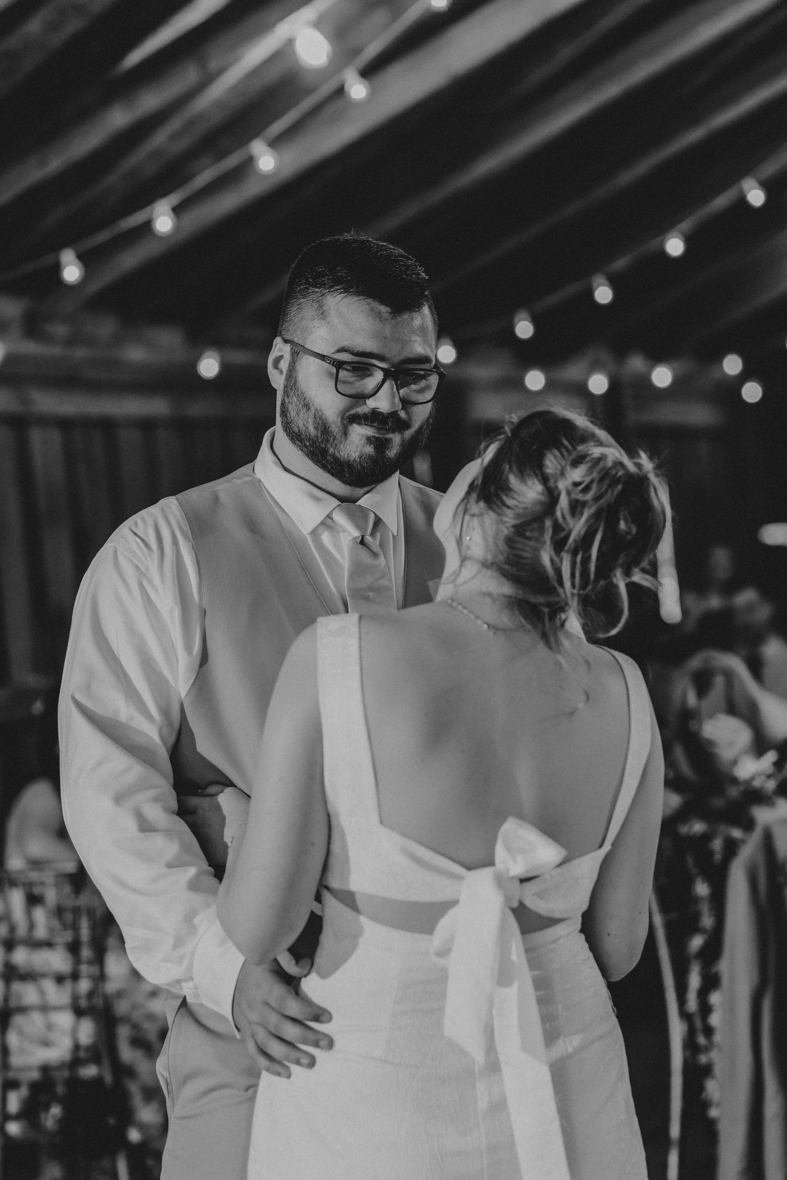 A couple embraces at a wedding reception under string lights, with the bride in a white dress and groom in a white shirt.