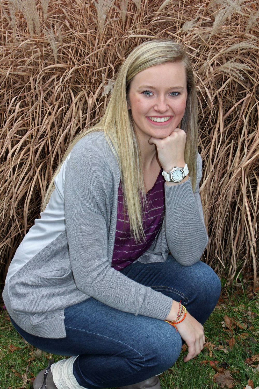 Smiling blonde woman crouching outdoors in jeans and gray cardigan, posing in front of tall dry ornamental grass.